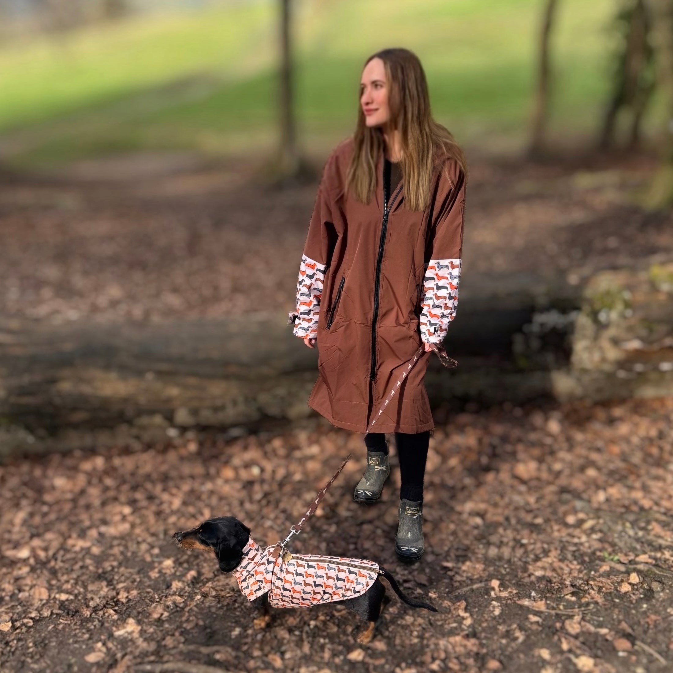 Woman wearing the DoxieBurrow waterproof dog walking robe coat with a dachshund wearing a matching coat outdoors.